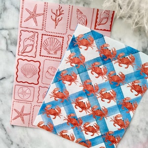 May include: Two sheets of decorative paper. One is pink with red seashell and starfish stamps. The other is blue and white checkered with red crab illustrations. The papers are likely for crafts or gift wrapping.