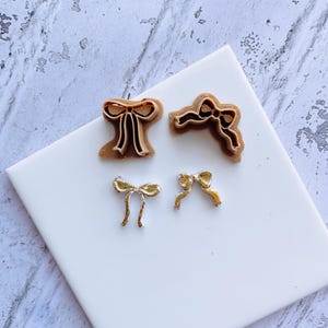 Puede incluir: Cuatro artículos en forma de lazo. Dos son cortadores de galletas de plástico marrón y dos son dijes de metal dorado. Los cortadores de galletas tienen forma de lazo con un diseño detallado. Los dijes son pequeños y delicados, con un acabado brillante.