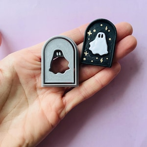 May include: A black and white cookie cutter and a black cookie with a white ghost design. The ghost has two eyes and a small mouth. The cookie is decorated with gold glitter.