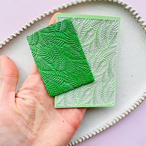 May include: Two green silicone embossing mats featuring a leaf design. The smaller mat is embossed with the pattern, while the larger mat is a stencil with the pattern cut out. The mats are on a white plate.