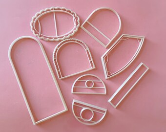 May include: A collection of white plastic cookie cutters in various geometric shapes, including ovals, arches, and rectangles, are arranged on a pink surface. The cutters have clean lines and are designed for baking.