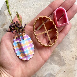 Puede incluir: Un pequeño jarrón de cuadros coloridos contiene ramitas de lavanda seca. También se ven un cortador de galletas con borde festoneado marrón claro y un cortador rosa en forma de arco. Los artículos se muestran sobre una superficie clara.