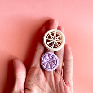 Può includere: Due timbri rotondi, decorativi, a forma di stella, in ceramica. Il timbro superiore è bianco con un motivo a stella in rilievo. Il timbro inferiore è viola con un motivo a stella in rilievo.