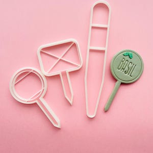 Może przedstawiać: Cztery białe plastikowe foremki do ciasteczek na różowym tle. Foremki mają kształt koła, prostokąta, długiego prostokąta i koła z napisem "Bazylia" w środku.