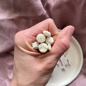 May include: A collection of white polymer clay stamps featuring various floral and geometric designs. The stamps are held in a hand and are ready to be used for crafting projects.