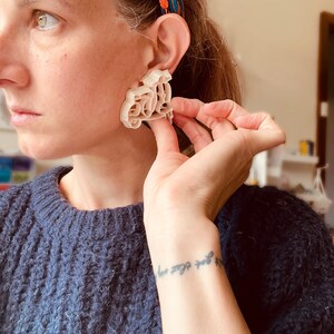 K&ouml;nnte beinhalten: Ein wei&szlig;er, 3D-gedruckter Ohrring in Form eines Rollers. Der Ohrring wird von einer Person mit einem blauen Pullover und einem Blumenkopfband getragen.