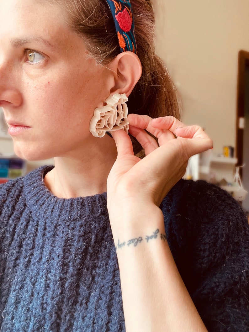 K&ouml;nnte beinhalten: Ein wei&szlig;er, 3D-gedruckter Ohrring in Form eines Rollers. Der Ohrring wird von einer Person mit einem blauen Pullover und einem Blumenkopfband getragen.