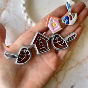 May include: A set of four cookie cutters featuring a birdhouse, two birds, and a bird with a house on its back. The cookie cutters are made of metal and are silver in colour.