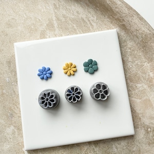 Puede incluir: Tres sellos de arcilla polimérica en forma de flor de color gris y tres pequeñas flores de arcilla polimérica en azul, amarillo y verde.