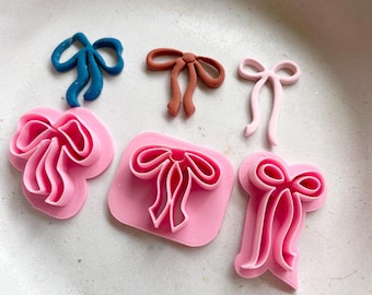 May include: A selection of bow-shaped craft tools in different colours. There's a teal bow, a brown bow, a light pink bow, and several pink bow-shaped cutters. The tools are arranged on a light pink surface.