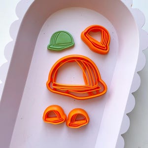 May include: A set of five cookie cutters in a light pink tray. The cutters are shaped like hats. There are four orange hat cutters and one green hat cutter.