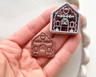May include: A brown gingerbread house cookie cutter with a heart detail and a white gingerbread house cookie cutter with a heart detail.