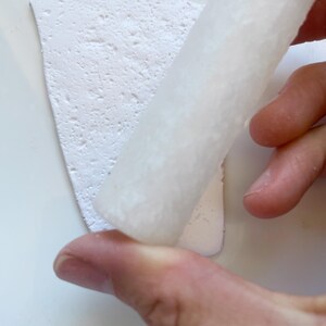 May include: A close-up of a white, textured clay slab being rolled with a white cylindrical tool. The clay has a rough, porous surface. The background is a plain white surface.