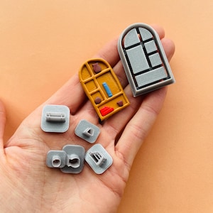 Może przedstawiać: Zestaw 3D-wydrukowanych foremek do ciasteczek w kolorze szarym i żółtym. Formy mają kształt okien, regału na książki i innych przedmiotów związanych z domem.