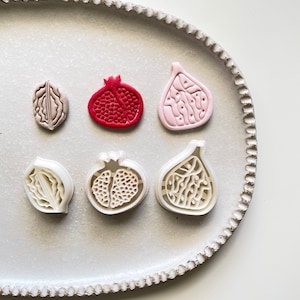 Op de afbeelding: Zes koekjesvormen op een wit bord. De vormen zijn in de vorm van een vijg, een granaatappel en een walnoot. De vijgvormen zijn roze en wit, de granaatappelvormen zijn rood en wit en de walnootvormen zijn bruin en wit.