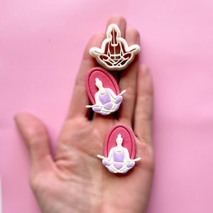May include: Three white plastic cookie cutters in the shape of a person sitting in a yoga pose. Two of the cutters are on a pink background and one is on a hand.