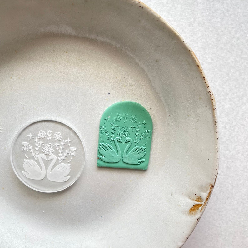 Puede incluir: Un sello de arcilla polim&eacute;rica verde con un dise&ntilde;o en relieve de dos cisnes que se miran con un dise&ntilde;o floral detr&aacute;s. El sello tiene forma de arco.
