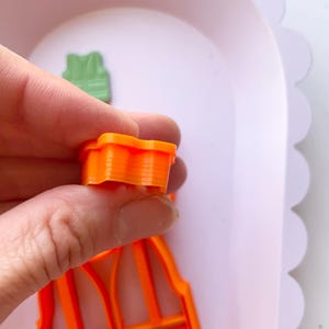May include: Two orange plastic cookie cutters shaped like a carrot. The cutter in the foreground is held in a hand. The cutter in the background is lying on a white surface.