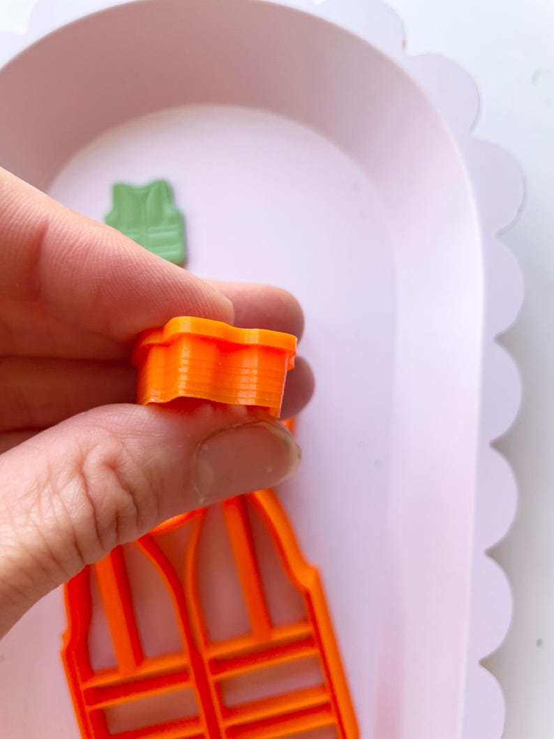 May include: Two orange plastic cookie cutters shaped like a carrot. The cutter in the foreground is held in a hand. The cutter in the background is lying on a white surface.