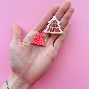 Op de afbeelding: Twee plastic kerstboomkoekjesvormen, een witte en een rode. De vormen worden in een hand tegen een roze achtergrond gehouden.