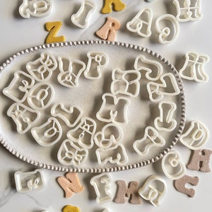 Peut inclure: Un ensemble d'emporte-pièces en plastique blanc en forme de lettres de l'alphabet, dans différents styles, disposés sur un plateau argenté avec des emporte-pièces supplémentaires éparpillés autour du plateau. Les emporte-pièces sont entourés de petites lettres en feutre de couleur.