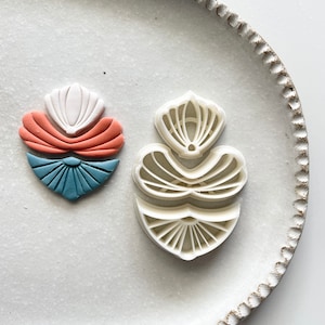 May include: Two white plastic cookie cutters in the shape of stylised flowers. The cutter on the left is layered with blue, orange and white fondant.