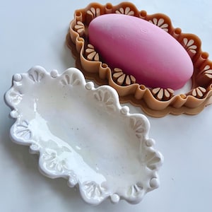 May include: A glossy white ceramic oval dish with a scalloped edge featuring embossed floral details, positioned next to a tan-coloured plastic mould containing a smooth, solid pink oval-shaped object that fits within the decorative frame.