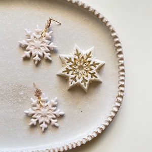 May include: Two white snowflake earrings with gold accents on a white plate. The earrings are made of polymer clay and have a delicate, intricate design.