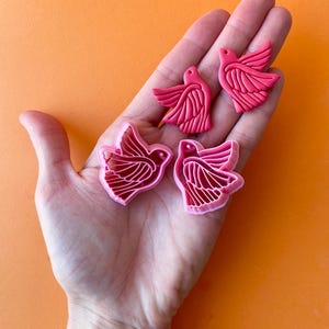 May include: Four pink and red clay bird earrings. The earrings are in the shape of a bird with detailed wings. The earrings are displayed on a hand.