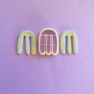 Puede incluir: Tres cortadores de galletas en forma de las letras "M-A-M" con un patrón a cuadros. Los cortadores son de color azul claro con un patrón a cuadros amarillos.