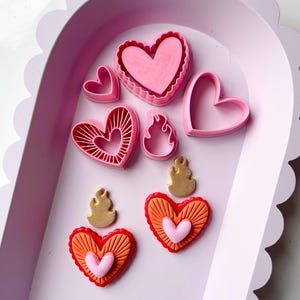 May include: A set of pink and red heart-shaped cookie cutters, including a large heart with a scalloped edge, a smaller heart with a scalloped edge, a heart with a sunburst design, and a heart with a flame design. There are also two flame-shaped cookie cutters.