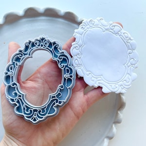 May include: Two gray plastic cake decorating molds with an ornate, decorative design. The molds are shaped like oval frames.