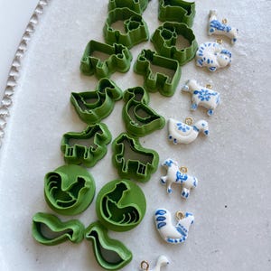Op de afbeelding: Een assortiment groene, diervormige koekjessnijders van plastic en kleine, witte keramische dierenbedels met blauwe bloemmotieven. De uitstekers bevatten koeien, paarden, eenden en hanen. De bedels hebben goudkleurige lussen.