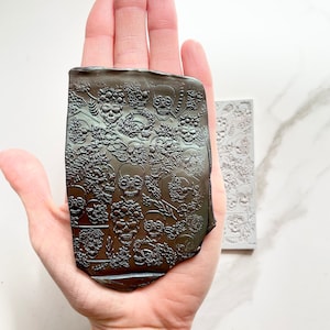 May include: A black silicone mold with a raised design of multiple skulls and floral patterns. The mold is being held in a hand.