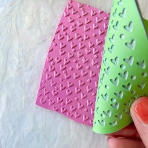 May include: A pink and green silicone stamp with a heart pattern. The pink stamp has a raised heart pattern and the green stamp has a cut-out heart pattern.