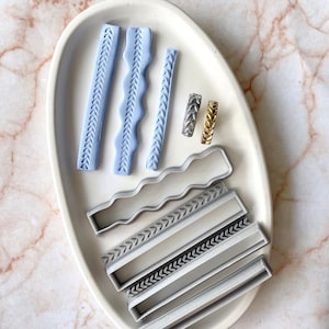 May include: A set of polymer clay cutters for creating decorative borders. The set includes four different designs: a wavy line, a chevron pattern, a straight line, and a leaf pattern. The cutters are arranged on a white plate with two gold and silver rings.