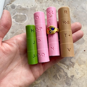Op de afbeelding: Een hand houdt vier cilindrische ringmaten in groen, roze en bruin. De roze maten hebben letters en cijfers, en een ervan bevat een gouden ring met rode en blauwe stenen. De ringmaten zijn voor het meten van ringmaten.