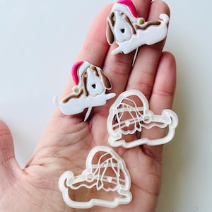 May include: Three white plastic cookie cutters shaped like a dog wearing a Santa hat. The cutters are on a person's hand.