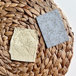 May include: Two floral patterned rubber stamps, one in a light yellow color and the other in a gray color, are displayed on a woven straw mat.