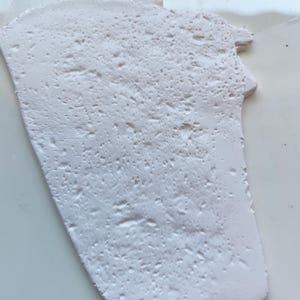 May include: A white, irregularly shaped piece of material with a textured surface. The surface has a pattern of small, raised bumps and ridges. The material appears to be solid and opaque, with a slightly uneven edge.