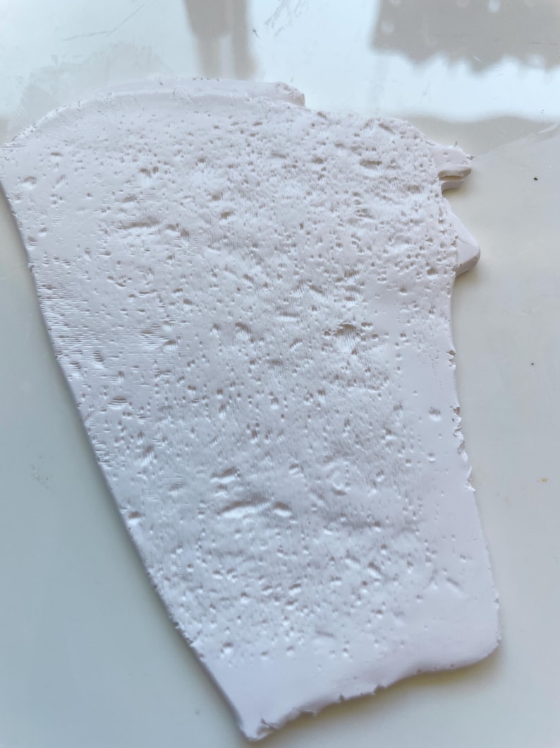 May include: A white, irregularly shaped piece of material with a textured surface. The surface has a pattern of small, raised bumps and ridges. The material appears to be solid and opaque, with a slightly uneven edge.