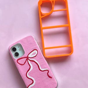 May include: Pink phone case with a hand-painted red and white bow design. An orange phone case frame sits next to the pink case. The background is a solid light pink.