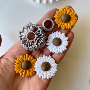 Op de afbeelding: Een set van vijf polymer clay-charms in de vorm van zonnebloemen en madeliefjes. De charms zijn in tinten geel, wit en bruin. De bloemen zijn allemaal ongeveer 2,5 cm in diameter.