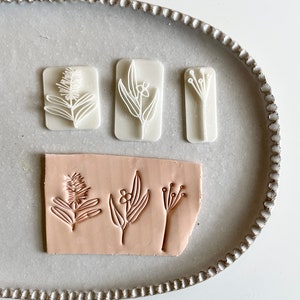 May include: Three white rubber stamps with floral designs on a light grey oval tray. The stamps are arranged in a row, and the designs are of a sprig of leaves, a flower with leaves, and a cluster of small flowers. A light pink piece of clay with the same floral designs stamped on it is in front of the stamps.