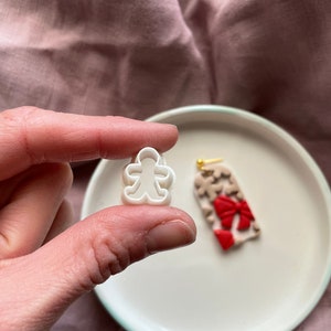 Pode incluir: Um cortador de biscoitos de gengibre de plástico branco em forma de homem. O cortador está sendo segurado em uma mão. No fundo, há um prato branco com um enfeite de gengibre vermelho e branco.