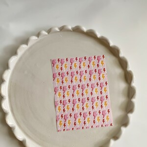 May include: A white ceramic plate with a scalloped edge, featuring a pink and yellow lightning bolt patterned paper on top.