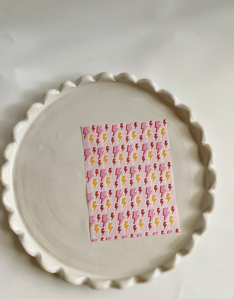 May include: A white ceramic plate with a scalloped edge, featuring a pink and yellow lightning bolt patterned paper on top.