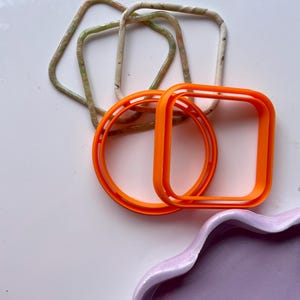 May include: A set of four bracelets, two are orange and two are white with green and brown marbling. The bracelets are all square shaped.