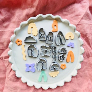 Pode incluir: Um prato de cerâmica branco com uma borda festonada, cheio de uma variedade de cortadores de biscoitos coloridos. Os cortadores têm a forma de nuvens, arco-íris, flores e outras formas abstratas.