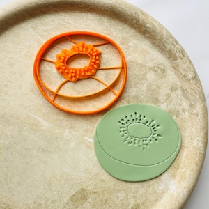 May include: An orange kiwi fruit cookie cutter and a green kiwi-shaped cookie. The cookie cutter is round with a detailed center and a circular outer ring. The green cookie has a detailed kiwi fruit design.